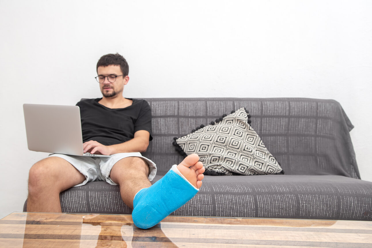 Man with glasses with broken leg blue splint treatment injuries from ankle sprain working laptop couch home |